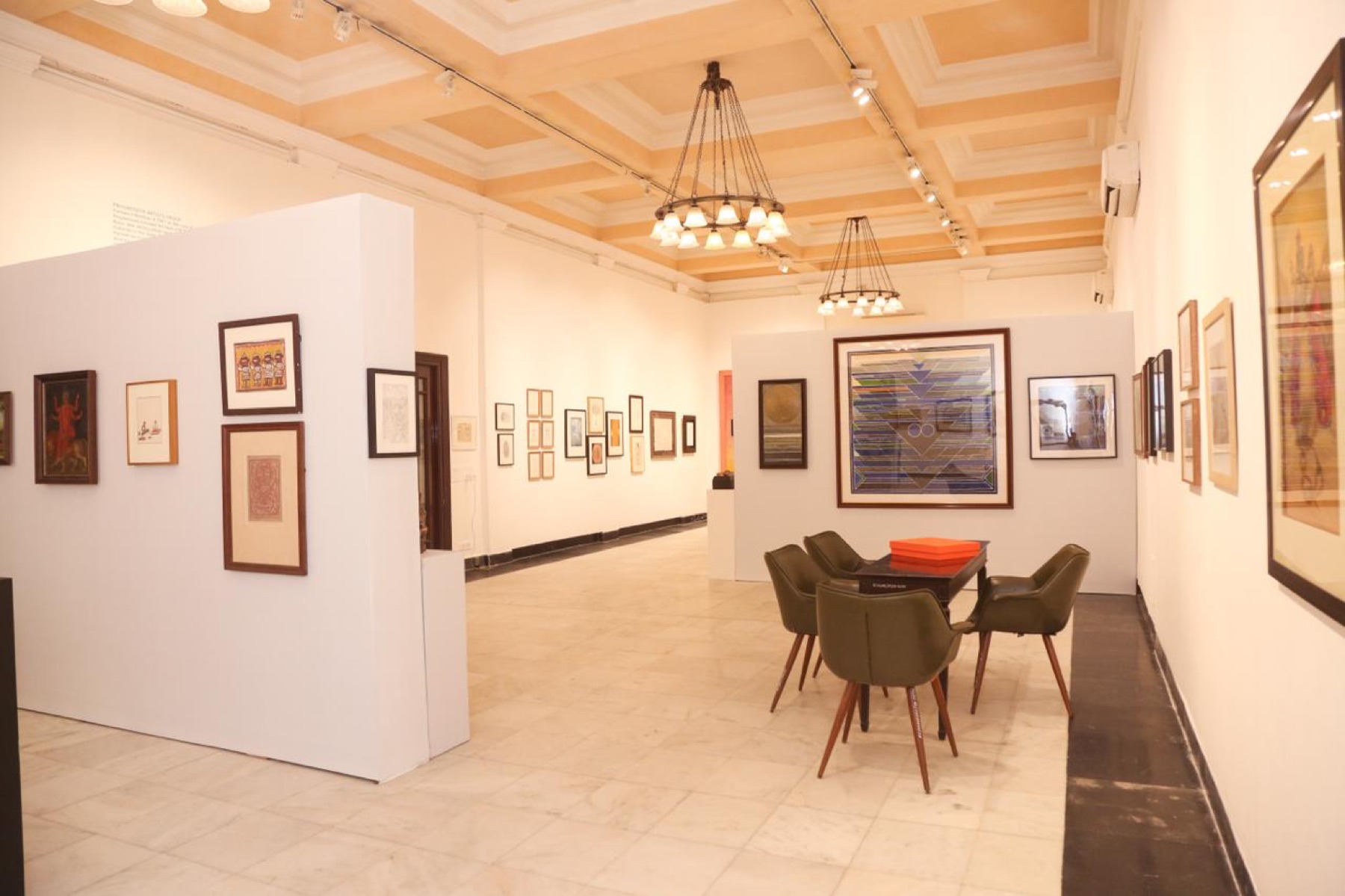 View through the exhibition: partition walls, paintings, and a study table with green chairs.